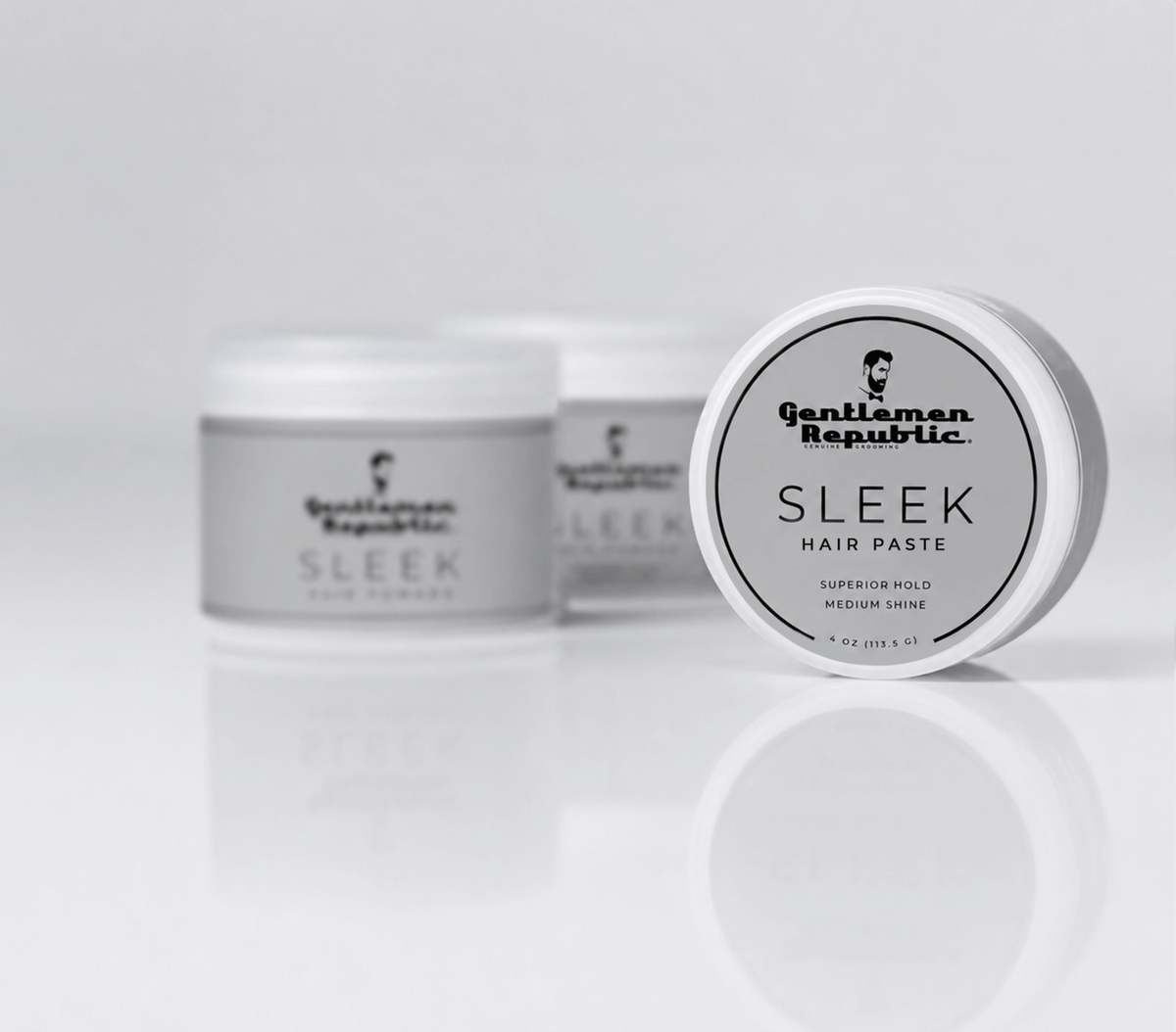 A round white plastic jar of Gentleman Republic Sleek Hair Paste in the Medium Shine variant, shown in sharp focus in the foreground with two more jars softly blurred in the background on a reflective white surface.