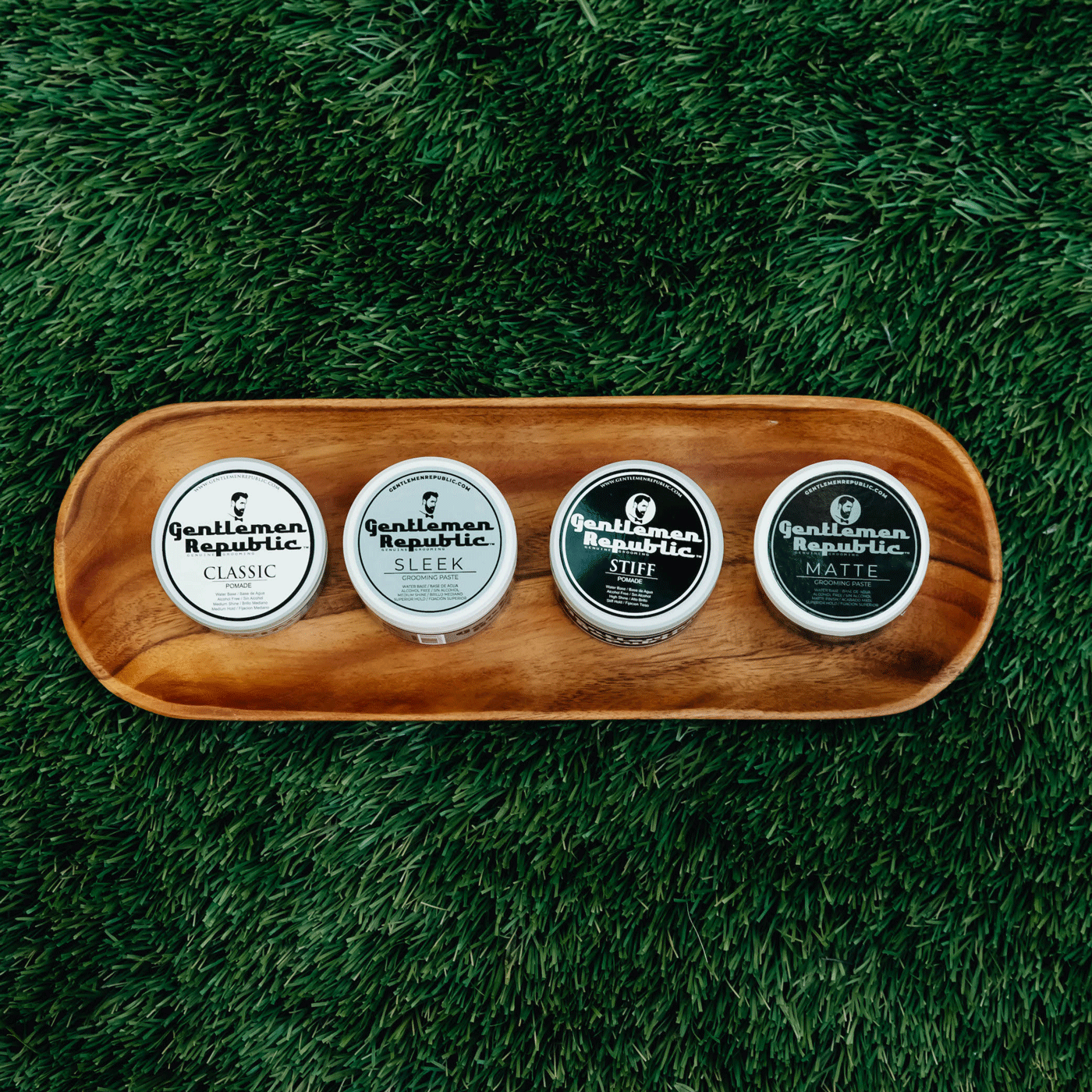 Four Gentlemen Republic pomades and pastes neatly arranged on a wooden tray against a background of lush green grass.